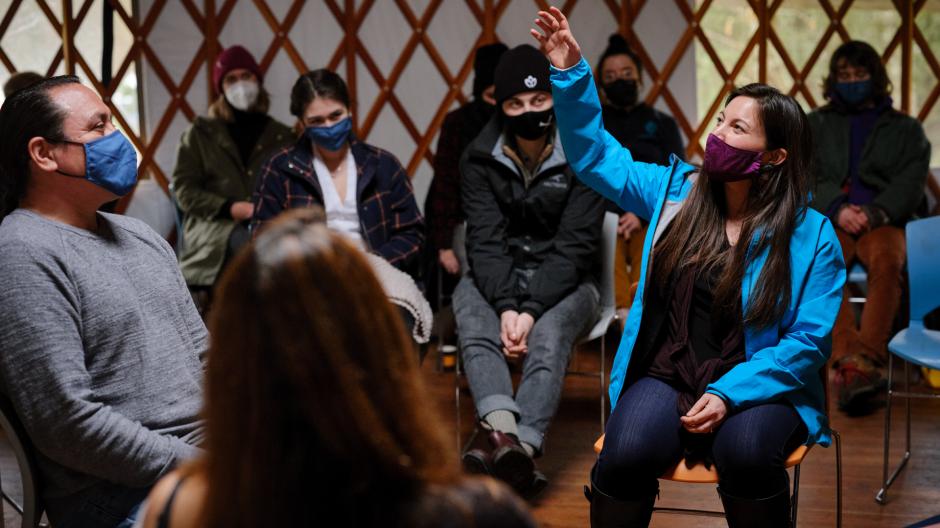 Group of people sitting inside of the UBC Farm yurt. One seated and speaking making hand gesture in direction of sky.