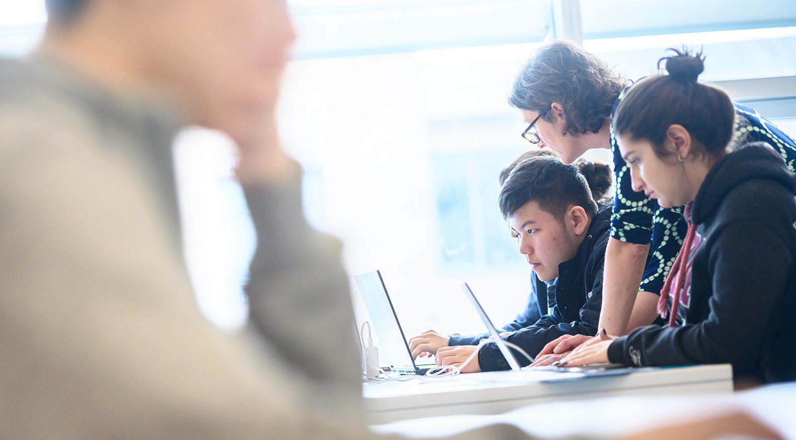 Professor instructing two students who are seated and on computers